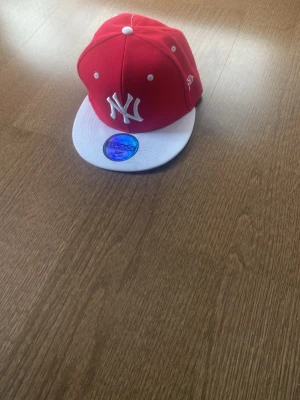 Röd New York Yankees snapback keps - Snygg röd snapback keps från New York Yankees med vit böjd skärm och broderad NY-logga framtill. Kepsen har klassisk snapback-stängning och är tillverkad i bomullsmaterial. Perfekt för dig som vill ha en sportig och trendig stil.