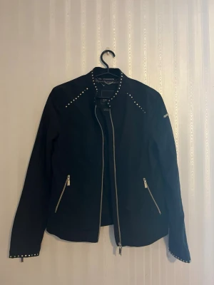 Svart vindjacka med nitar - Svart vindjacka från GEOX med guld nitar längs axlar, rygg och ärmsluy. Jackan har dragkedjor framtill, på fickorna och vid ärmsluten.