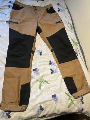 Bruna cargobyxor med svarta detaljer - Säljer ett par bruna cargobyxor med svarta förstärkningar på knän, bak och fickor. Byxorna har flera dragkedjefickor och resår vid benslut. Perfekta för dig som gillar funktionella och snygga byxor med streetkänsla.