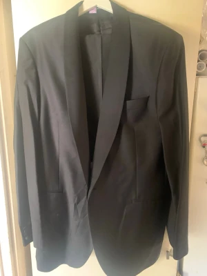 Svart smoking  från Oscar Jacobson - Snygg svart smoking  från Oscar Jacobson med klassisk sjalkrage och slät design. Kavajen har två framfickor och en bröstficka, perfekt för att höja stilen. Materialet är slätt och ger en elegant känsla.