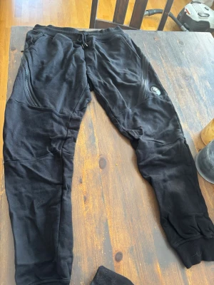 Svarta mjukisbyxor från C.P. Company - Säljer ett par svarta mjukisbyxor från C.P. Company med coola detaljer som dragkedjor på fickorna och den ikoniska linsen på ena benet. Byxorna har ribbade muddar vid benslut och är gjorda i mjukt material som är perfekt för chill dagar.