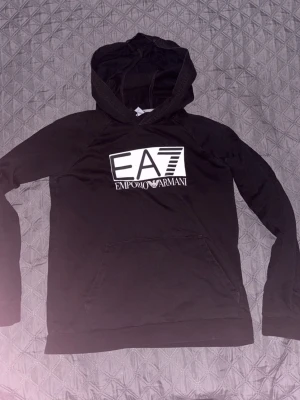 Svart hoodie EA7 Emporio Armani - Snygg svart hoodie från EA7 Emporio Armani med stor vit logga på bröstet. Klassisk huva och känguruficka framtill. Tillverkad i mjukt material som känns skönt mot huden. Perfekt för en avslappnad och trendig look.