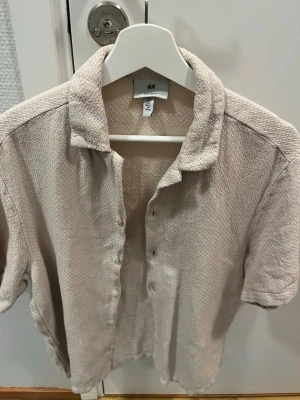 Beige kortärmad skjorta från H&M - Snygg beige skjorta från H&M i ett mjukt, strukturerat material. Skjortan har korta ärmar, klassisk krage och knappar framtill. Perfekt för en avslappnad och stilren look.