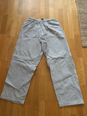 Grå mjukisbyxor från Collusion - Säljer ett par grå mjukisbyxor från Collusion Studios med tryck på vänster sida. Byxorna har resår i midjan och raka ben. Perfekta för chill dagar och har en avslappnad passform. Materialet är mjukt och skönt, troligtvis bomull eller bomullsblandning.