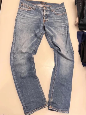 Blå jeans från Nudie Jeans - Säljer ett par klassiska blå jeans från Nudie Jeans med raka ben och fem fickor. Jeansen har snygga slitningar och kontrastsömmar samt Nudie-loggan på bakfickan. Tillverkade i mjuk denim som ger en avslappnad look. Passar som 32/33. Väldigt flexibel med priset