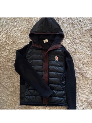 Marinblå  Moncler cardigan - Säljer en svart cardigan från Moncler med huva och vinröda detaljer. Jackan är väldigt sällsynt och nästan svårt att få tag på då den är OG. Den har inga defekter och använts skonsamt vid frågor och funderingar så är det bara att skriva. Självklart äkta. Jackan sitter dessutom perfekt ifall du har storleken S och sitter inte för löst och piffigt på kroppen