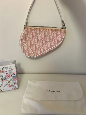 Christian Dior rosa Saddle Bag - Ikonisk Christian Dior Saddle Bag i rosa och vitt med monogrammönster. Köpt i Paris 2004. Väskan har en unik sadelform, vit axelrem och dragkedja upptill. Tillverkad i canvas med detaljer i vitt läder. Perfekt statement piece för dig som vill sticka ut.