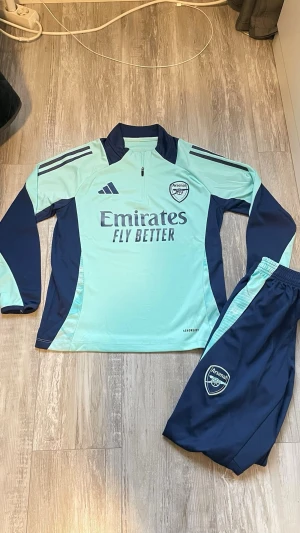 Arsenal FC Training Set 24/25 – Adidas - Säljer nu ett komplett Arsenal träningsställ från Adidas i en riktigt fräsch mintgrön/marinblå färgkombination. Setet inkluderar zip träningströja och tillhörande träningsbyxor. Mycket skönt material med AEROREADY-teknologi. 