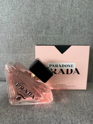 Prada Paradoxe Eau de Parfum - Prada Paradoxe Eau de Parfum i en unik trekantig glasflaska med ljusrosa doftvätska och svart fyrkantigt lock. Flaskan har tydlig Prada-logga och kommer med en stilren rosa kartong. Perfekt för dig som gillar exklusiva och moderna parfymer.