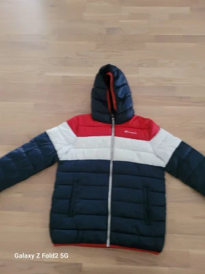Champion pufferjacka marinblå/röd XL - Snygg pufferjacka från Champion i marinblått, vitt och rött. Jackan har huva, dragkedja framtill och två sidofickor. Perfekt för kalla dagar med sin vadderade design och sportiga look. Passar dig som gillar streetwear och vill hålla värmen.