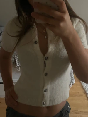 Vit kortärmad kofta med pärlknappar - Söt vit kofta med korta ärmar och pärlknappar framtill. Modellen är croppad och har ribbade kanter vid ärmslut och nederkant. Perfekt att styla med höga jeans eller kjol för en trendig look.