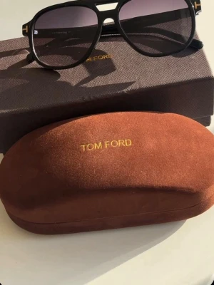 Svarta solglasögon från Tom Ford - Snygga svarta solglasögon från Tom Ford med klassisk aviatorform och mörka glas. Bågarna är i svart syntetmaterial med gulddetaljer vid sidorna. Kommer med originalbrun sammetsfodral och ask, båda med Tom Ford-logga i guld.