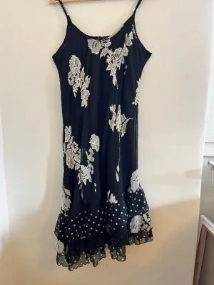 Svart blommig klänning med volang - Svart midiklänning med vita blommor och prickar. Tunna axelband och volangkant nertill ger en luftig och feminin look. Perfekt för sommarens varma dagar. Klänningen är snygg och fräsch. 