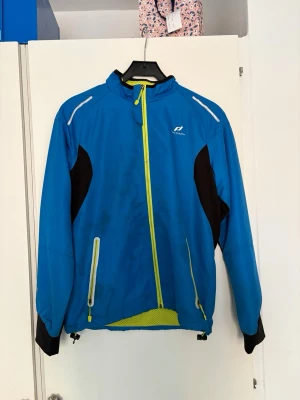 Blå vindjacka från Pro Touch - Sportig blå vindjacka från Pro Touch med svarta paneler på sidorna och gula dragkedjor. Jackan har hög krage, två fickor med dragkedja och reflekterande detaljer på axlarna. Perfekt för utomhusaktiviteter när det blåser.