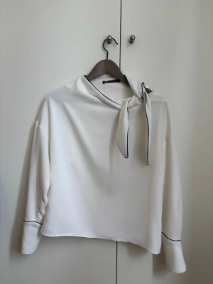 Vit blus med knytband från Zara - Stilren vit blus från Zara med knytband i halsen och svarta kontrastsömmar. Blusen har långa ärmar och en rak passform, perfekt för en trendig och chic look.