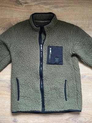 Olivgrön teddyjacka med dragkedja - Olivgrön teddyjacka från KappAhl i 100% polyester med hel dragkedja framtill, svarta detaljer och en bröstficka med dragkedja och tryckt text. Två sidofickor med svarta kanter och hög krage för extra mysfaktor. Perfekt för kalla dagar och riktigt snygg till jeans.