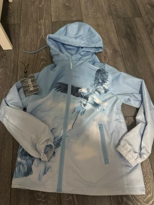Ljusblå vindjacka med örntryck - Snygg ljusblå vindjacka med huva och dragkedja framtill. Jackan har ett stort tryck av flygande örnar i blå och vita nyanser på framsidan. Två fickor med dragkedja och elastiska muddar vid ärmsluten. Perfekt för dig som vill sticka ut med en unik design.