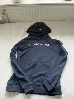 Mörkblå hoodie från Ocean Works - Snygg mörkblå hoodie från Ocean Works med vit logga på bröstet och liten logga på huvan. Klassisk känguruficka framtill och ribbade muddar vid ärmar och nederkant. Tillverkad i mjukt bomullsmaterial och har dragsko i huvan.