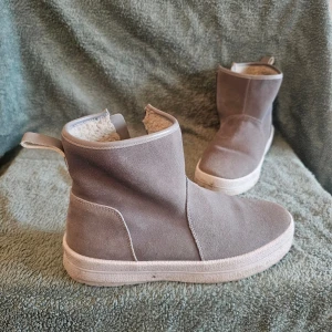 Gråa mockastövletter med foder - Snygga gråa stövletter i mocka med dragkedja på sidan och mjukt, ljust teddyfoder inuti. Skorna har en rund tå, vit sula och en enkel, stilren design. Perfekta för dig som vill ha både stil och komfort under kalla dagar.