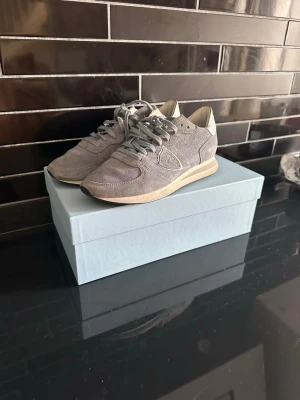 Grå Philippe Model sneakers i mocka - Snygga grå sneakers från Philippe Model i mocka med vita detaljer och klassisk låg siluett. Skorna har en vit sula och diskret logga på sidan. Perfekta för dig som gillar stilrena och trendiga sneakers med premiumkänsla.