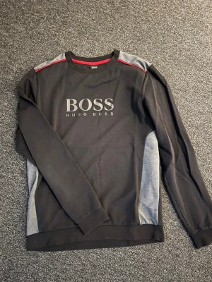 Svart långärmad tröja från Hugo Boss - Snygg svart långärmad tröja från Hugo Boss med grå paneler på sidorna och axlarna samt röda detaljer vid axlarna. Stor BOSS-logga framtill. Perfekt för en clean och sportig look.