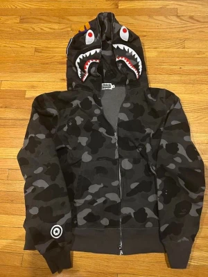 Svart camo hoodie med hajmotiv - Säljer en svart camo zip-hoodie från A Bathing Ape med hajmotiv på huvan och röda detaljer. Hoodien har dragkedja framtill, känguruficka och ribbade muddar. Perfekt för dig som gillar streetwear och vill sticka ut med en unik design.