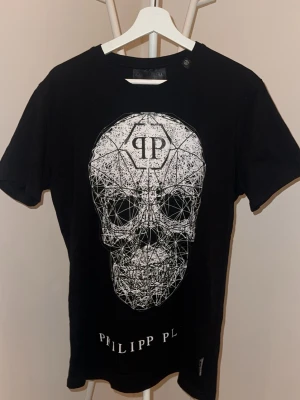 Svart t-shirt med döskalle från Philipp Plein - Cool svart t-shirt från Philipp Plein med stor vit grafisk döskalle på framsidan och logga. Klassisk passform med rund hals och korta ärmar. Perfekt för dig som gillar edgy streetstyle och vill sticka ut med en unik design.