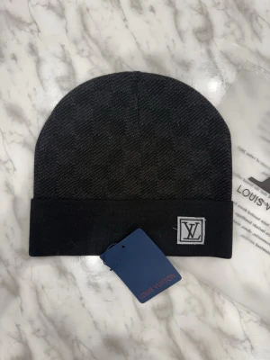 En mörk svart mössa  - En mörk svart stickad mössa med rutigt mönster och uppvikt kant, med en liten logotyp framtill och etikett kvar.