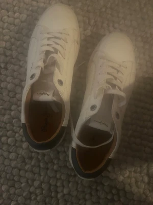 Vita sneakers från Pepe Jeans - Snygga vita sneakers från Pepe Jeans med klassisk design. Skorna har vita snören, diskret logga på sidan och mörk blå detalj vid hälen. Tillverkade i syntetmaterial med bekväm innersula. Perfekta för en stilren och enkel look. Andvänt fåtal gånger tex som runt 5 nypris 800kr