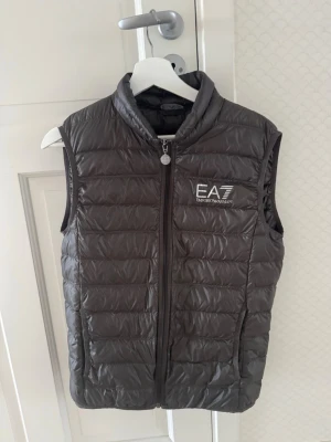 Mörk grön dunväst från EA7 Emporio Armani - Snygg svart dunväst från EA7 Emporio Armani med diskret glansig finish och logga på bröstet. Västen har hög krage, dragkedja framtill och två sidofickor. Perfekt att slänga över en hoodie eller tröja för en sportig och trendig look.