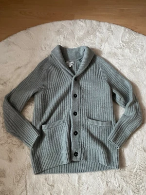 Grå ribbstickad kofta med sjalkrage - Mysig grå ribbstickad kofta med sjalkrage och svarta knappar framtill. Två stora fickor fram och långa ärmar. Perfekt för lager på lager och chill stil. Materialet är mjukt och värmande, ullblandning. Storleken är S, sprillans ny aldrig andvänd utomhus.