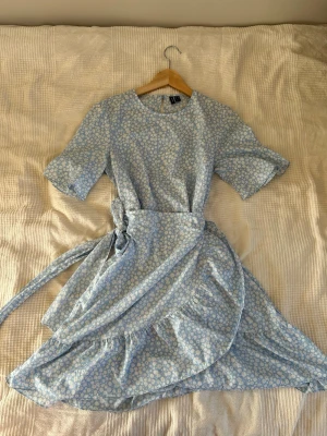 Blommig ljusblå omlottklänning Vero Moda - Supersöt ljusblå omlottklänning från Vero Moda med vitt blommönster. Klänningen har korta ärmar, volangkant nertill och knytband i midjan för en snygg passform. Perfekt för sommarens alla tillfällen! Endast använd 1 gång💗