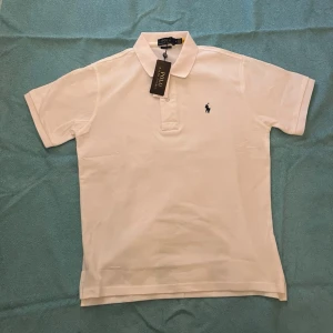 Vit pikétröja från Polo Ralph Lauren - Klassisk vit pikétröja från Polo Ralph Lauren med korta ärmar och krage. Tröjan har den ikoniska broderade loggan på bröstet och är tillverkad i mjuk bomull. Perfekt till jeans eller shorts för en clean och stilren look.