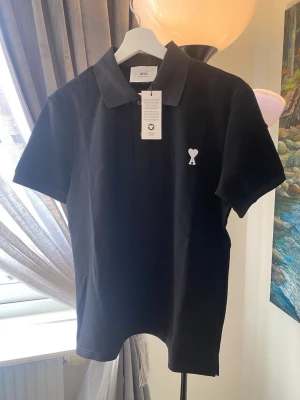 Svart pikétröja från Ami Paris - Klassisk svart pikétröja från Ami Paris med korta ärmar och vit broderad logga med hjärta på bröstet. Tröjan har krage och knappar framtill. Perfekt till en clean och stilren look.