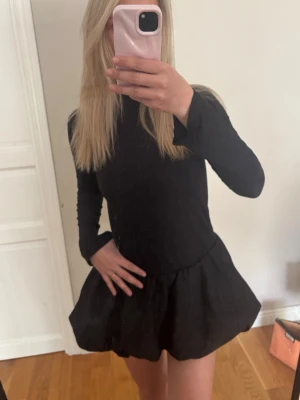 Svart kort klänning med puffkjol - En svart kort klänning med långärmad design och volang nedtill. Klänningen har en enkel och stilren look. Perfekt för dig som gillar minimalistisk stil och vill ha något som sticker ut med sin volangdetalj.  Jag är 165❣️