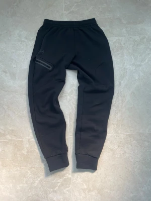 Svarta mjukisbyxor från Under Armour - Säljer ett par svarta mjukisbyxor från Under Armour med elastisk midja och ribbade benslut. Byxorna har en snygg detalj med en dragkedjeförsedd ficka på sidan och diskret logga. Perfekta för chill eller träning, tillverkade i mjukt material. Storlek: S. Skicket är extremt fint. Pris går att diskutera☺️