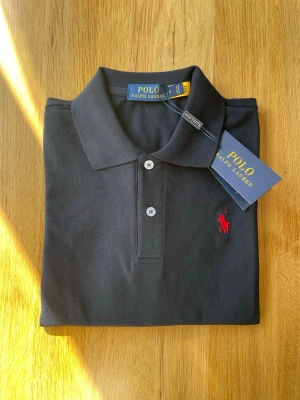 Svart pikétröja från Polo Ralph Lauren - Marinblå pikétröja från Polo Ralph Lauren med klassisk krage och två knappar framtill. Tröjan har det ikoniska röda broderade logotypen på bröstet och är tillverkad i ett mjukt bomullsmaterial. Perfekt för en stilren och avslappnad look.