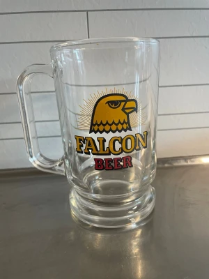 Falcon Beer ölglas med örntryck - Stort ölglas i transparent glas med tryck av en gul örn och texten 'Falcon Beer' i svart och rött. Glaset har ett rejält handtag och tjock botten, perfekt för att njuta av en kall dryck.