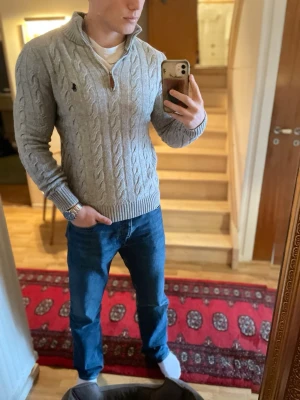 Grå kabelstickad tröja Polo Ralph Lauren - Snygg grå kabelstickad tröja från Polo Ralph Lauren med halv dragkedja och hög krage. Tröjan har ett broderat logotyp på bröstet och ribbade muddar vid ärmslut och nederkant. Perfekt för lager-på-lager och en klassisk look.