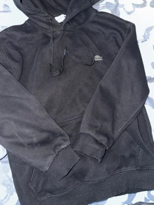 Svart hoodie från Lacoste - Svart hoodie från Lacoste med klassisk krokodillogga på bröstet. Hoodien har dragsko i huvan, magficka och ribbade muddar vid ärmslut och nederkant. Perfekt för en avslappnad och stilren look.