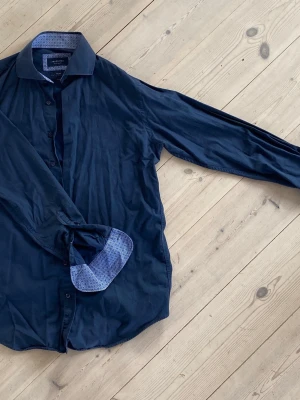 Mörkblå skjorta från Profuomo - Snygg mörkblå skjorta från Profuomo med lång ärm och klassisk krage. Insidan av krage och manschetterna har ett diskret mönster i ljusblått. Skjortan har knappar framtill och är tillverkad i mjuk bomull. Modell: Slim fit. Storlek: 39.