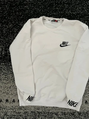 Vit sweatshirt från Nike - Säljer en stilren vit sweatshirt från Nike med svart logga på bröstet och tryckt Nike-text på muddar vid ärmslut. Tröjan har rund halsringning och är tillverkad i mjukt material, perfekt för en avslappnad och sportig look.