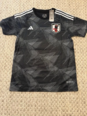 Japan svart matchtröja Adidas - Snygg svart matchtröja från Adidas med Japans landslagsmärke på bröstet. Tröjan har vita detaljer, geometriskt mönster och klassiska Adidas-ränder på axlarna. Tillverkad i lätt och ventilerande material, perfekt för träning eller match.