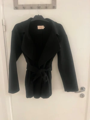 Svart kappa med knytskärp från Nelly - Stilren svart kappa från Nelly med breda slag och knytskärp i midjan. Kappan har en rak passform och är tillverkad i ett mjukt material som ger en snygg siluett. Perfekt för att höja din outfit under kyliga dagar. Säljer den på grund av att den är för stort och därför är den även knappt använd, bara några få gånger, och har inga slitningar alls 