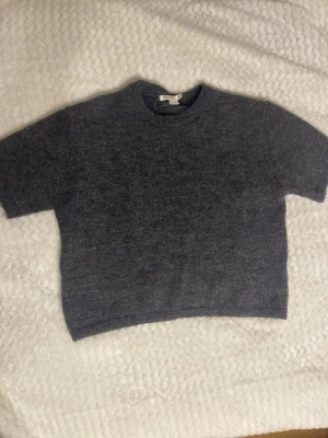 Stickad tröja kortärmad - Mysig mörkgrå stickad tröja med korta ärmar och rund halsringning. Tröjan har en enkel och stilren design, perfekt att matcha med jeans eller kjol. Materialet ser ut att vara mjukt och värmande, perfekt för kyligare dagar.