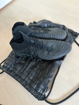 Nike phantom luna elite (kolla beskrivning) - Säljer ett par svarta Nike fotbollsskor med hög socka och snörning. Passar 45,5 men är 46. Skorna har mönstrad ovandel i syntetmaterial och AG-dobbar  för bra grepp på planen. Nike-logga i turkos på sulan. Perfekta för dig som vill ha stil och funktion på matchen. Köp gärna med instabox för att få den skickad samma dag (innan kl 16)!