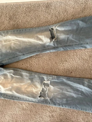 Ljusblå slitna jeans med hål - Säljer ett par ljusblå jeans från Nelly med slitningar och stora hål på båda benen. Jeansen har en unik ficka med lock och knappdetalj. Perfekta för dig som gillar en avslappnad och trendig stil.