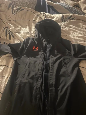 Svart vindjacka från Under Armour - Snygg svart vindjacka från Under Armour med huva och dragkedja framtill. Jackan har en röd logga på bröstet och texten UNDER ARMOUR längs ryggen. Tillverkad i ett lätt och vindtätt material, perfekt för blåsiga dagar. Ord pris 1200 men säljs för 750