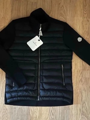 Moncler Cardigan - Snygg svart dunjacka från Moncler med quiltad front och släta ärmar. Jackan har två dragkedjefickor fram, hög krage och Moncler-logga på ärmen. Perfekt för kalla vinterdagar och ger en stilren look.