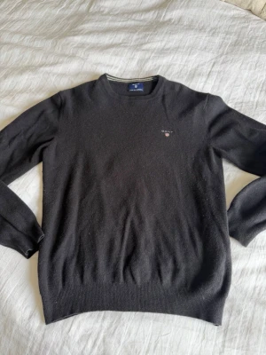 Svart stickad tröja från Gant - Klassisk svart stickad tröja från Gant i mjuk lammull. Tröjan har rund halsringning, ribbade muddar och en liten broderad Gant-logga på bröstet. Perfekt för dig som gillar stilrena och tidlösa plagg.